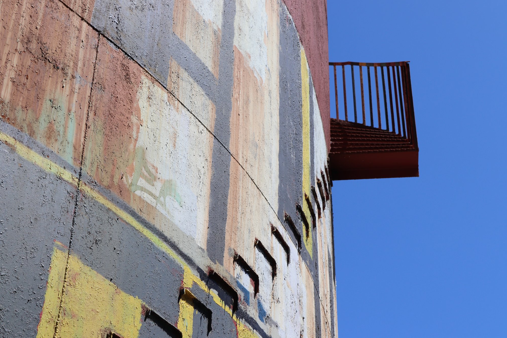 Weathered painted concrete structure with a rusted balcony against a blue sky