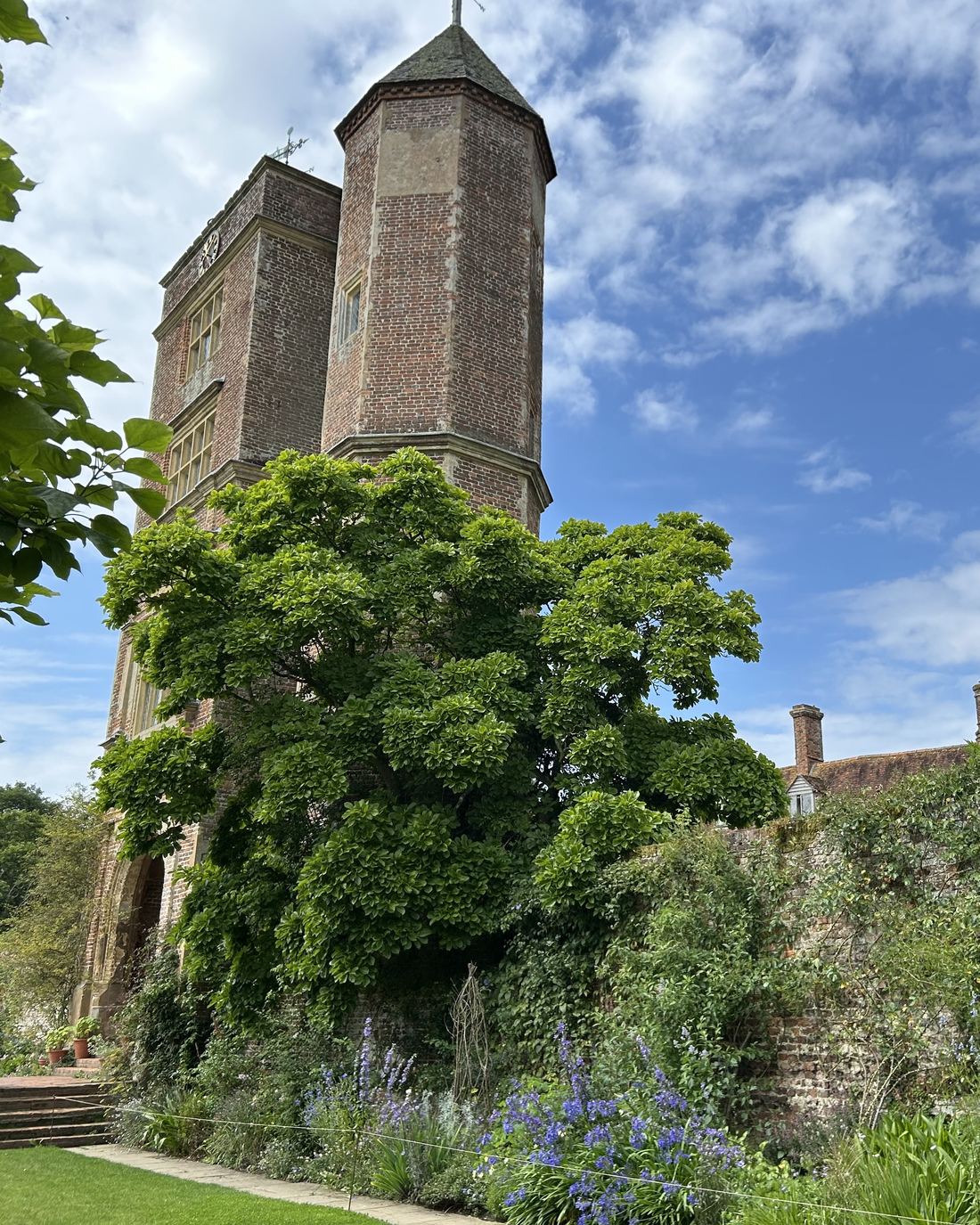 Historic brick tower with mullioned windows framed by a garden wall, climbing greenery and flowering border